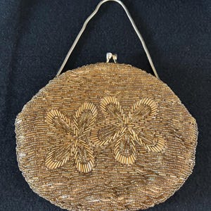 May include: A vintage gold beaded handbag with a silver chain handle. The round purse features two large floral designs made of vertical beads. The bag has a clasp closure and is set against a dark background.