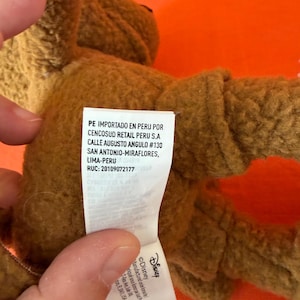 May include: A brown plush toy with a tag that includes the Disney logo and text. The tag also includes text in Spanish. The toy is on an orange background.