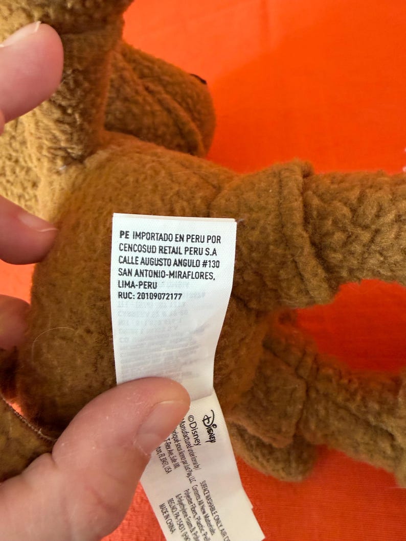 May include: A brown plush toy with a tag that includes the Disney logo and text. The tag also includes text in Spanish. The toy is on an orange background.
