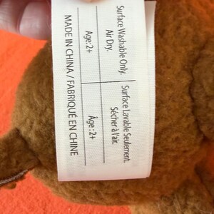 May include: A brown plush toy with a tag that reads "MADE IN CHINA / FABRIQU&Eacute; EN CHINE". The tag also states "Surface Washable Only" and "Air Dry". The toy is recommended for ages 2+.
