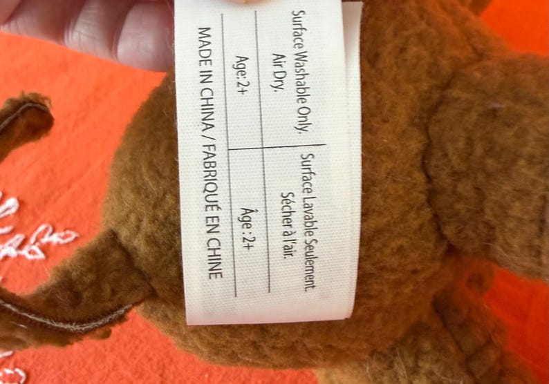 May include: A brown plush toy with a tag that reads "MADE IN CHINA / FABRIQU&Eacute; EN CHINE". The tag also states "Surface Washable Only" and "Air Dry". The toy is recommended for ages 2+.