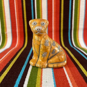 May include: A ceramic figurine of a sitting leopard with an orange and yellow glaze. The leopard has gray spots and is set against a colorful striped background. The figurine is a decorative item.
