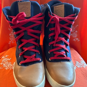 Op de afbeelding: Paar hoge Reebok Classic sneakers. De schoenen hebben een marineblauwe bovenkant met bruine leren neuzen en witte zolen. Rode veters lopen door de marineblauwe bovenkant. Het Reebok-logo is zichtbaar op de tong.
