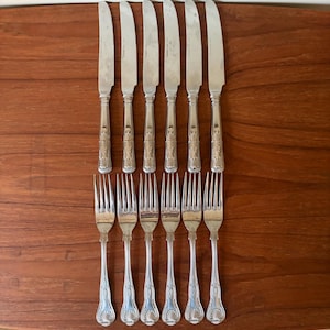 May include: A set of six silver-toned metal dinner knives and six matching dinner forks arranged in two rows. Each piece features a polished finish and a decorative embossed shell motif at the base of the handle, with slender, tapered stems.