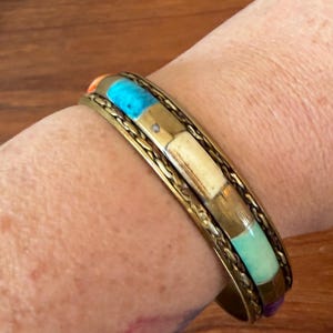 May include: A brass cuff bracelet featuring a central row of rectangular inlaid stones in shades of turquoise, cream, and light green, bordered by two twisted metal wire bands. The bracelet has a metallic finish and a smooth, open-ended design.
