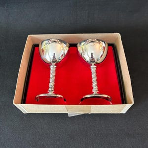 May include: A pair of silver goblets with decorative stems, displayed in a box with a red velvet interior. The goblets have a reflective surface, and the stems have a twisted pattern. The box is cardboard.