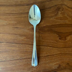 Puede incluir: Una cuchara de metal plateado con un acabado liso y reflectante y un mango cónico. El cuenco de la cuchara tiene forma ovalada y el mango presenta un sutil diseño de cresta vertical. La palabra STAINLESS está claramente grabada en el cuenco.