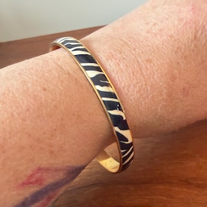 Op de afbeelding: Een stijve armband met een contrastrijk zebrastrepenpatroon in zwarte en crèmekleurige emaille. De ronde band is afgewerkt met een gepolijste goudkleurige metalen rand langs beide zijden, wat zorgt voor een strak en gedefinieerd profiel.