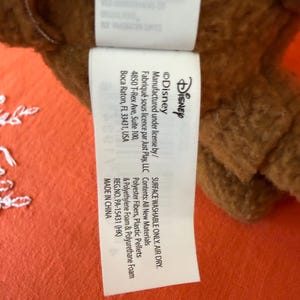 May include: A close-up of a white tag with black text, attached to a brown plush toy. The tag features the Disney logo and mentions the materials used, including polyester fibres and plastic pellets. The tag also states the product is surface washable and air dry.