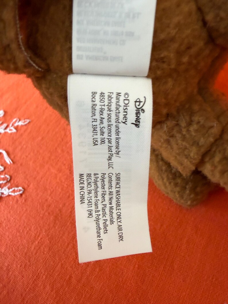 May include: A close-up of a white tag with black text, attached to a brown plush toy. The tag features the Disney logo and mentions the materials used, including polyester fibres and plastic pellets. The tag also states the product is surface washable and air dry.