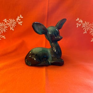 以下が含まれることがあります： 横たわったポーズのセラミック製の鹿の置物。鹿は光沢のある濃いティール色で、黒のアクセントが施されています。置物は明るいオレンジ色の背景に置かれています。