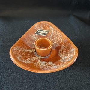Puede incluir: Un portavelas de cerámica marrón con un glaseado blanco. El portavelas tiene una forma triangular y una sola copa para velas en el centro. El portavelas tiene una etiqueta que dice "Wilton Pottery, Burlington, Canada".