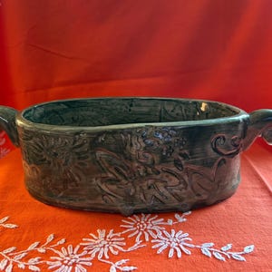 Puede incluir: Una fuente de cerámica verde oscuro con patrones florales y abstractos en relieve. La fuente ovalada tiene dos asas curvas y se encuentra sobre una superficie naranja brillante.