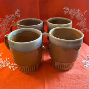 May include: Four ceramic coffee mugs with a stacked design. The mugs have a brown base, a light blue and white rim, and a brown handle. The mugs are on an orange tablecloth with white floral embroidery.