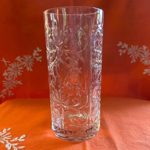 May include: A clear, tall glass with an intricate floral pattern etched into the surface. The glass is cylindrical and appears to be made of crystal or high-quality glass. The background is a vibrant orange fabric.