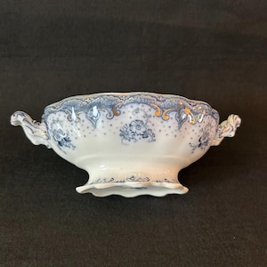 Puede incluir: Un cuenco de porcelana blanco y azul con motivos florales y detalles dorados. El cuenco tiene dos asas y un borde y base festoneados. El diseño presenta motivos florales azules y ribetes dorados.