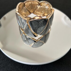 May include: A gold-toned metal bracelet with black and gray triangular enamel segments. The bracelet is resting on a reflective surface, showcasing its design and color contrast. The bracelet's design is geometric and modern.