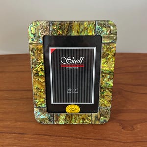 May include: Rectangular photo frame with a shimmering, iridescent shell-like border. The frame has a black interior with the word "Shell" and the dimensions 3.5" x 5" (9cm x 13cm) printed on it. The frame is sitting on a wooden surface.