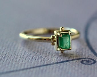 May include: A gold ring featuring a rectangular emerald gemstone. The emerald is set in a square bezel with decorative accents. The ring band is thin and polished, creating a classic and elegant look.