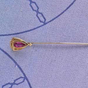 May include: A gold stick pin featuring a pear-shaped, deep pink gemstone set within a geometric, triangular gold frame. The pin has a long, slender, straight metal needle extending from the decorative head, showcasing a polished metallic finish.