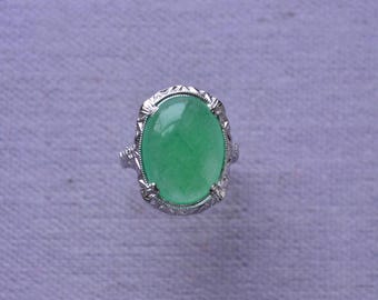 May include: A silver ring featuring a large, oval-shaped, translucent green gemstone. The ring has an ornate, filigree-style setting with detailed patterns around the gemstone. The ring is set against a light purple background.