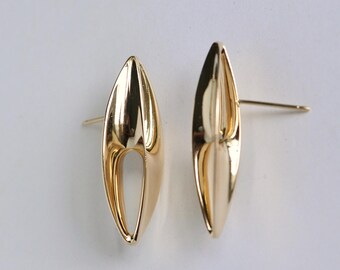 May include: A pair of gold-toned earrings with a unique, abstract design. The earrings are shaped like a long, thin oval with a curved opening in the center.