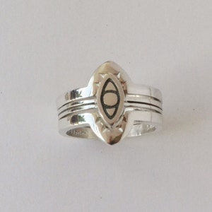 May include: A silver ring with a stylized eye design in the center. The ring has a wide band with three grooves.