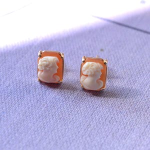 May include: A pair of gold tone earrings with cameo insets. The cameos are rectangular and feature a white cameo profile of a woman with a delicate design.