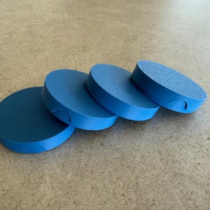 May include: Four blue, round coasters are arranged diagonally across the frame. Each coaster has a smooth, solid blue top and a textured, slightly lighter blue side. A small notch is cut into the side of each coaster.