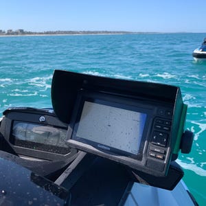 May include: A black and white Garmin GPS navigation device with a sunshade mounted on a black and white jet ski. The jet ski is on the water with a blue sky and a shoreline in the background.