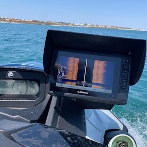 Puede incluir: Un buscador de peces y una unidad de visualización Garmin negros montados en una embarcación. La pantalla muestra datos de sonar con una profundidad de 7,8 m. El salpicadero muestra la velocidad y otra información. El fondo es un océano azul con una costa distante.