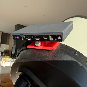 Può includere: Un dispositivo elettronico nero con più porte, montato su una staffa rossa, attaccato a un grande telescopio cilindrico. Il dispositivo ha porte HDMI, USB e altre. Il telescopio è color crema.