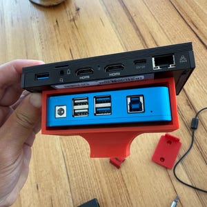 Può includere: Un dispositivo nero con più porte, tra cui HDMI, USB ed Ethernet, è montato su un supporto rosso stampato in 3D. Il dispositivo è collegato a un dispositivo blu con quattro porte USB.