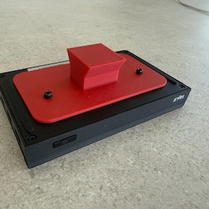 Può includere: Un dispositivo elettronico nero con una parte superiore rettangolare rossa e un componente rosso a forma di cubo. Due viti nere fissano la parte superiore rossa. Il dispositivo ha un pulsante di accensione e il testo "MULE" è visibile.