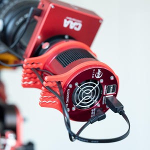 Puede incluir: Primer plano de una cámara roja y negra. La cámara tiene un cuerpo cilíndrico con un ventilador y puertos USB. El conjunto está montado sobre una estructura de soporte roja. Cables negros conectados.