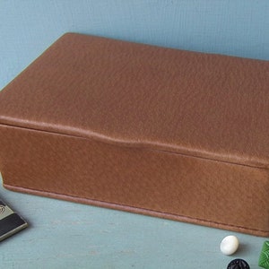 May include: A rectangular brown box with a textured surface. The box has a slightly curved top and is likely made of leather or a similar material. A small card and several buttons are visible nearby.