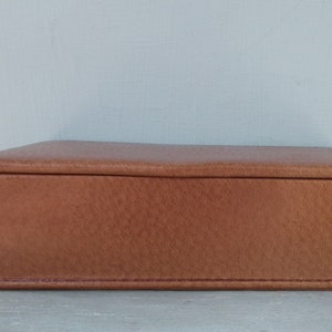 May include: A rectangular, brown leather box with a textured surface. The box has a hinged lid and is likely used for storage. The box is sitting on a light gray surface.
