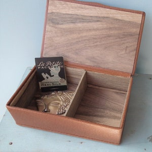 May include: Open, rectangular, brown leather box with a wooden interior and a divider. Inside the box is a small card with the text "La Petite Paquette" and a decorative metal face. The box is open, revealing the interior.