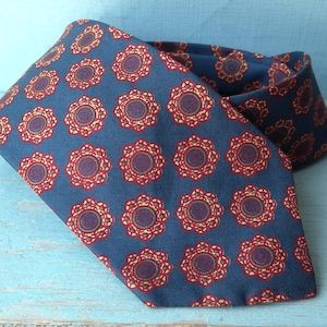 May include: A blue necktie featuring a repeating pattern of circular floral motifs in shades of red, gold, and dark blue. The fabric has a visible diagonal weave texture, and the tie is folded to display the pointed end and the neckband section.