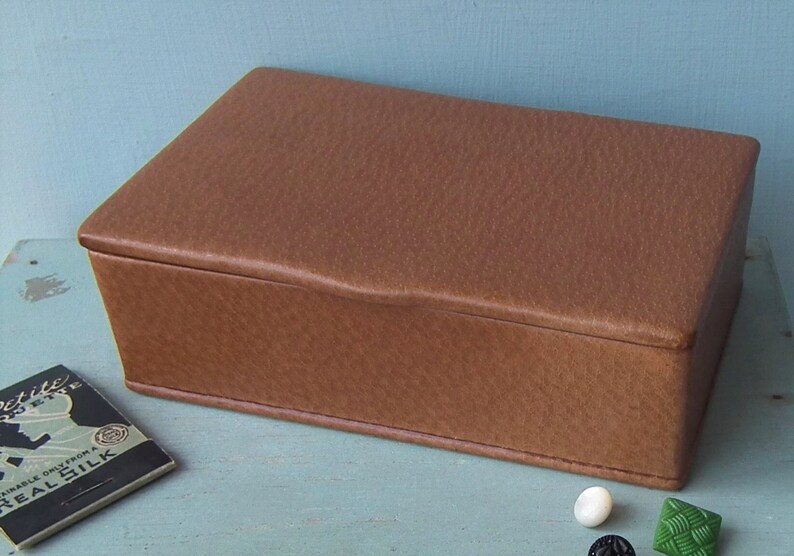 May include: A rectangular brown box with a textured surface. The box has a slightly curved top and is likely made of leather or a similar material. A small card and several buttons are visible nearby.