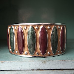May include: A wide, silver-toned cuff bracelet with a decorative design. The bracelet features alternating oval-shaped inlays of deep red and green, set against a gold-toned background. Small silver studs accent the edges.