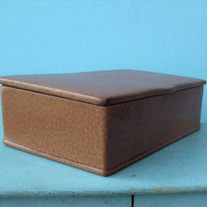 May include: A rectangular, brown leather box with a textured surface and a slightly curved lid. The box is set against a light blue background, showcasing its simple, classic design. The box is likely used for storage.