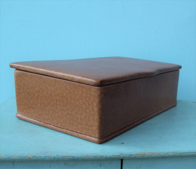 May include: A rectangular, brown leather box with a textured surface and a slightly curved lid. The box is set against a light blue background, showcasing its simple, classic design. The box is likely used for storage.