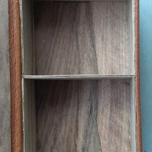 May include: Open, rectangular wooden box with a light brown leather exterior. The interior is divided into two compartments by a horizontal shelf. The wood grain is visible inside the box.