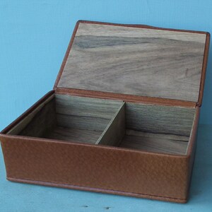 May include: Open rectangular box with a hinged lid. The exterior is brown leather, and the interior is divided into two compartments with a wooden base and lid. The box is designed for storage.