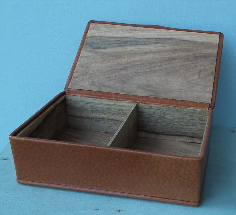 May include: Open rectangular box with a hinged lid. The exterior is brown leather, and the interior is divided into two compartments with a wooden base and lid. The box is designed for storage.