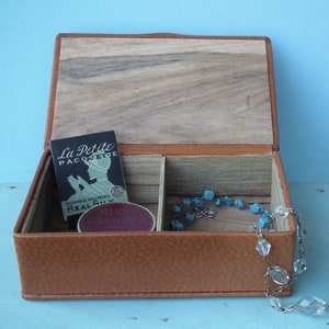 May include: Open brown leather jewelry box with a wooden lid and interior. Inside are two compartments, a small card with text, and a turquoise beaded necklace. A clear crystal necklace hangs over the side.