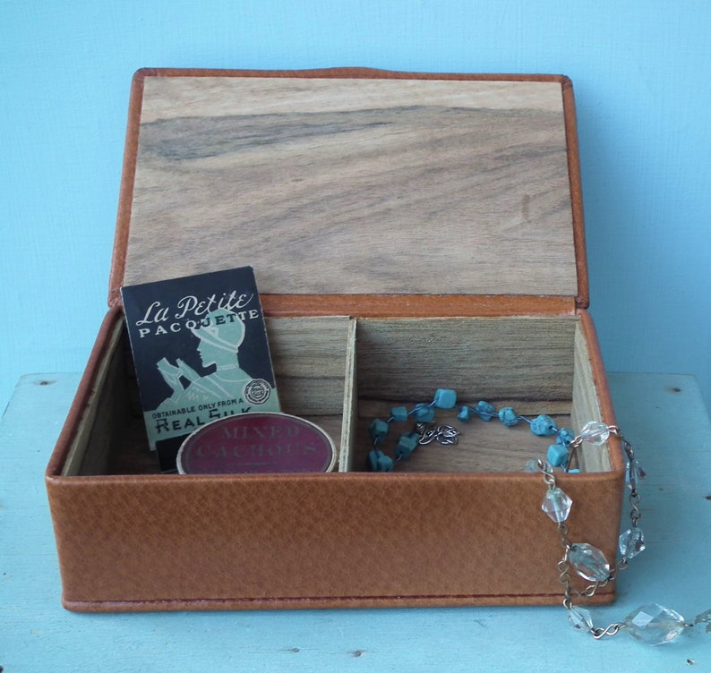 May include: Open brown leather jewelry box with a wooden lid and interior. Inside are two compartments, a small card with text, and a turquoise beaded necklace. A clear crystal necklace hangs over the side.