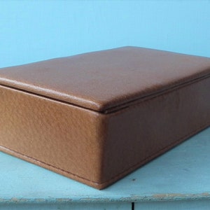 May include: A rectangular brown leather box with a lid. The box is a light brown color and appears to be made of leather. The box is sitting on a light blue surface.