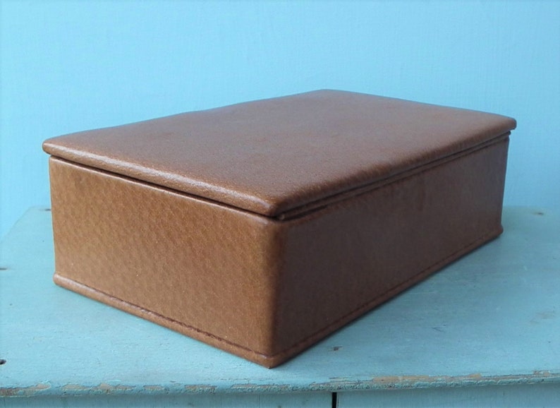 May include: A rectangular brown leather box with a lid. The box is a light brown color and appears to be made of leather. The box is sitting on a light blue surface.